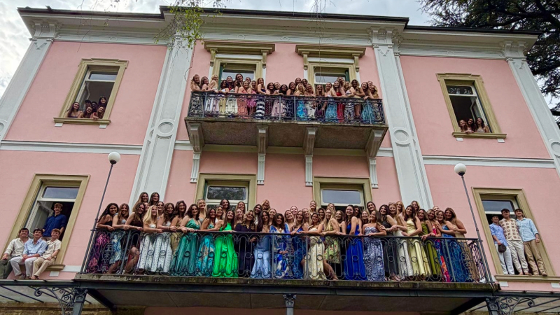 Students stand on balconies and in windows of a pink multi-story building, the Montarina Hostel in Lugano.