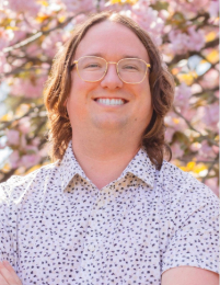 Logan Hammerschmitt stands in front of a pink tree wearing a white shirt specked with black dots.