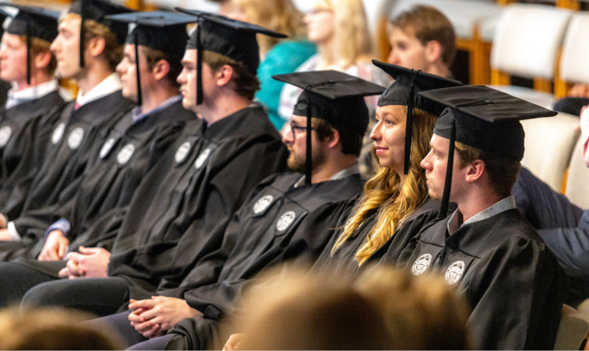 A row of students in black graduation caps and gowns sits in seats in Antrim Chapel and they are listening intently.