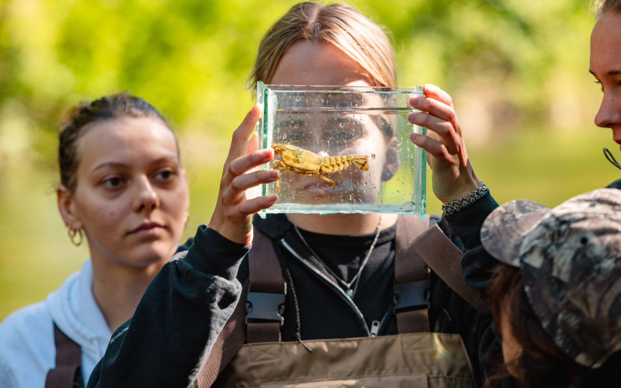 A student holds a clear container filled with water and a rust-colored crawdad in front of her face. Three other students visible in the frame look at the crawdad with her. 