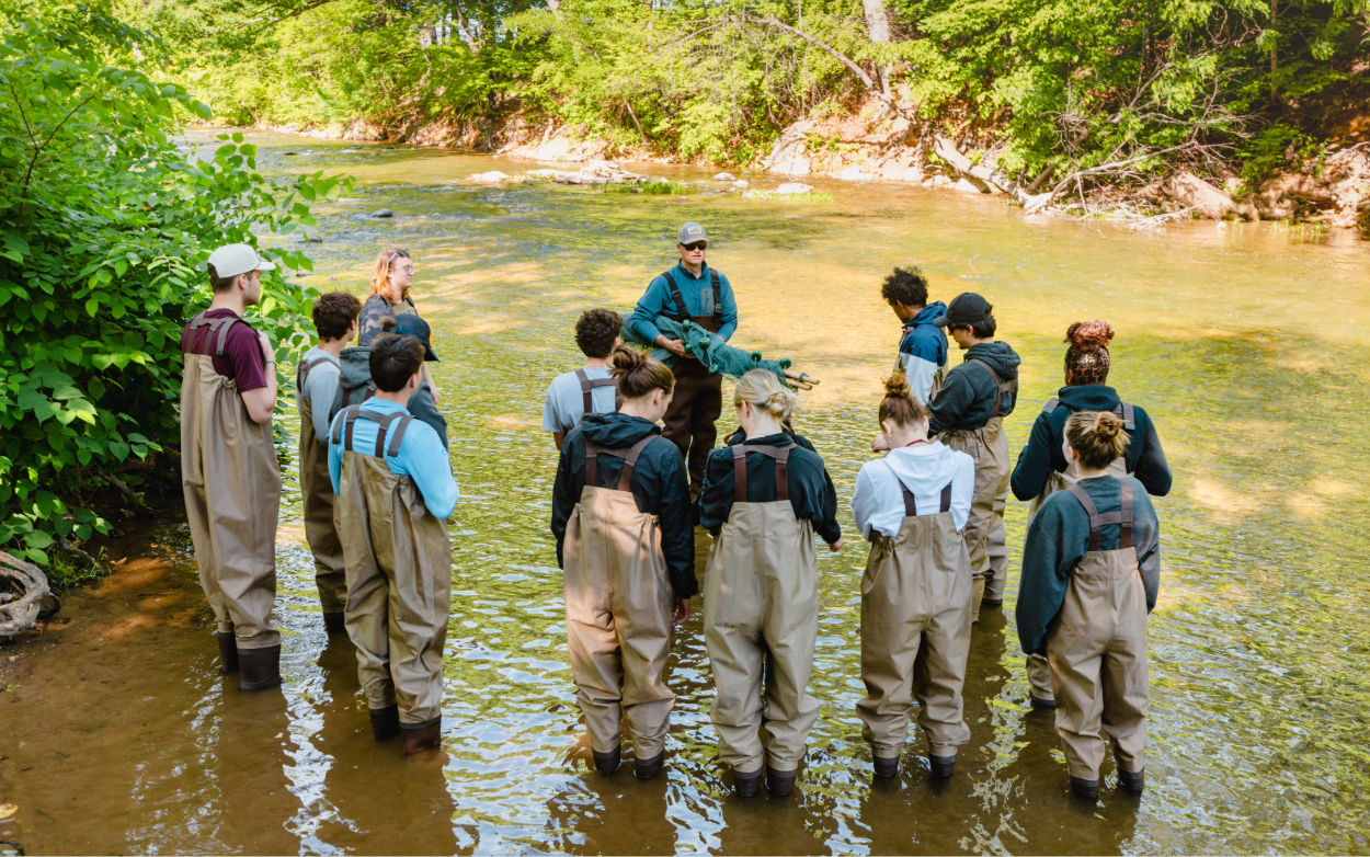 Steve Powers speaks to a group of students in the river. They are all wearing waders. Steve wears a hoodie under his waders, a hat and sunglasses, and is holding a large rolled-up green net. 
