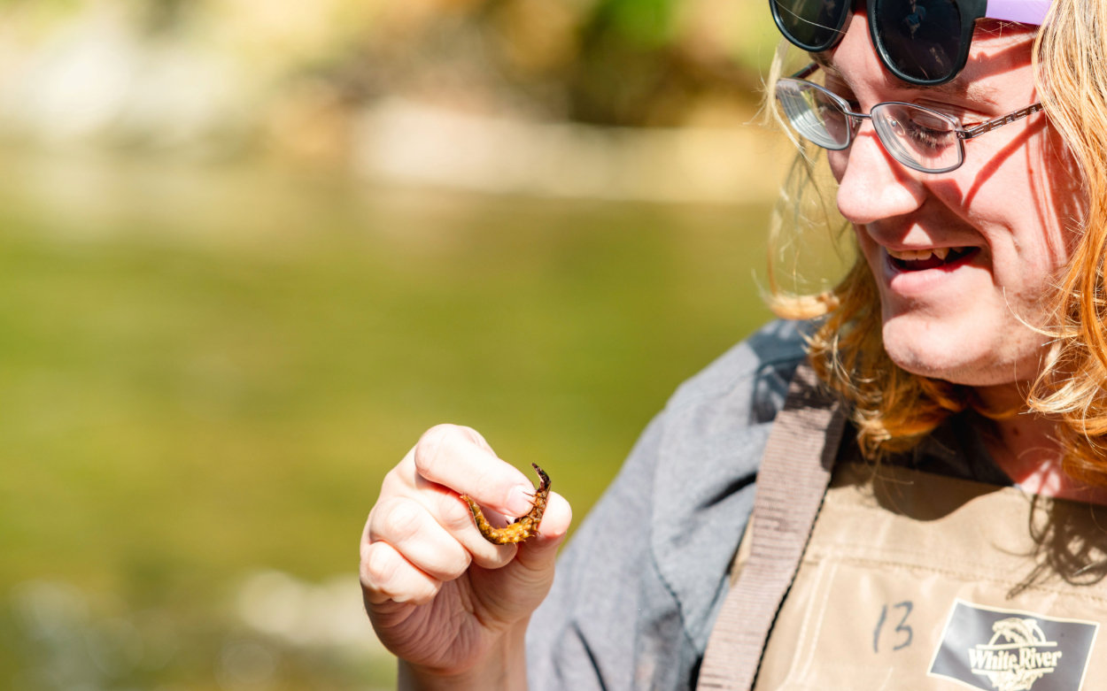 A student with shoulder-length light hair smiles as they hold a small river bug of sorts. The bug is long, brownish-yellow, has lots of legs and is curled up just slightly.  