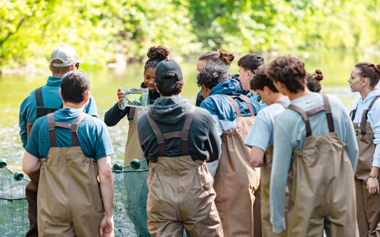 A group of people stand in their waders in the river. They are gathered around one student, who is holding a clear container full of water and fish.