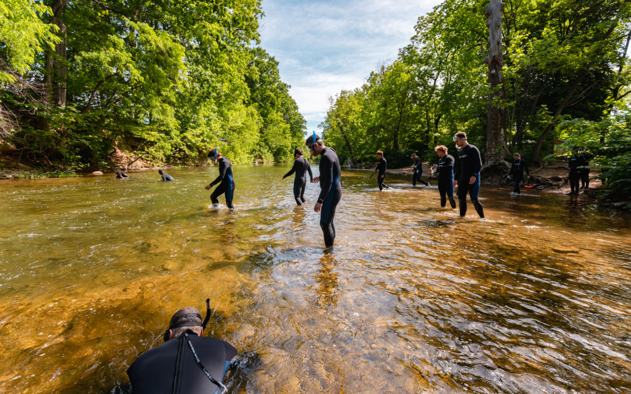 Someone in the foreground snorkels in shallow water in the river. Other people are scattered around, standing in the shallow water in their wetsuits. 