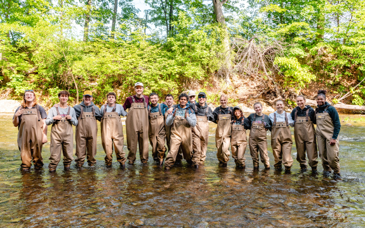 Sixteen students stand in their waders in the river and pose for a group shot. The trees behind them are very green. One student in the center holds up one of the clear containers. 