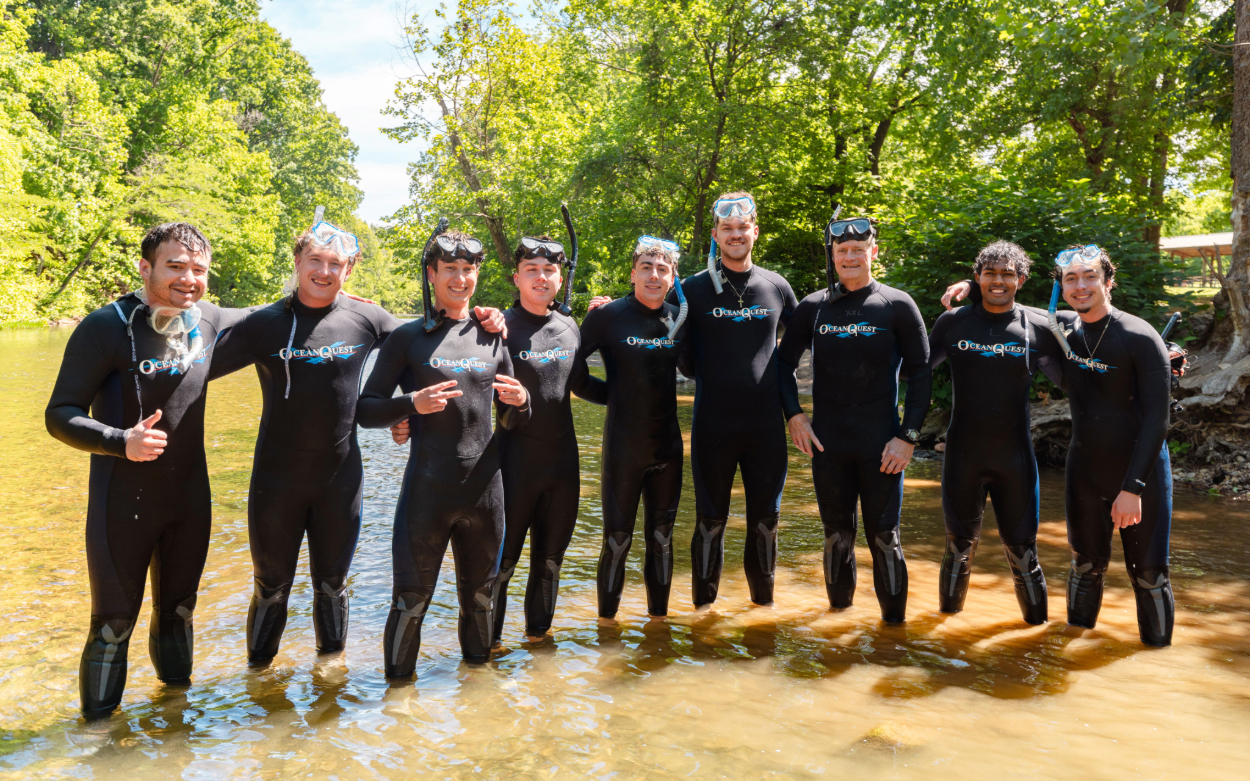 Nine men stand in the river and pose for a photo. They are all wearing black wetsuits, and all of them have snorkels and goggles either in their hands, on top of their heads or around their necks. 