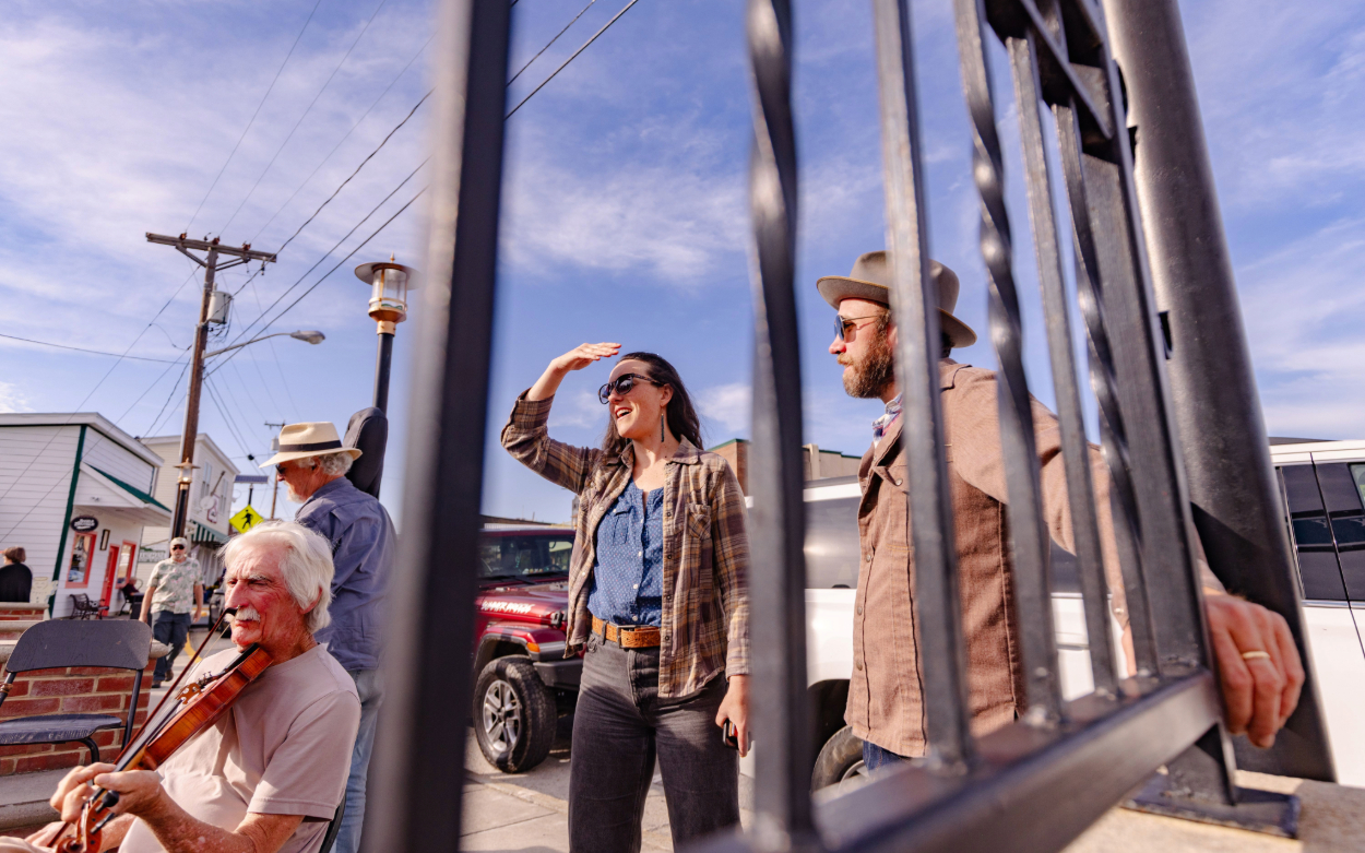 Two people stand looking off into the distance, slightly obscured by wrought-iron bars. An older man sits to their left and plays the fiddle. 