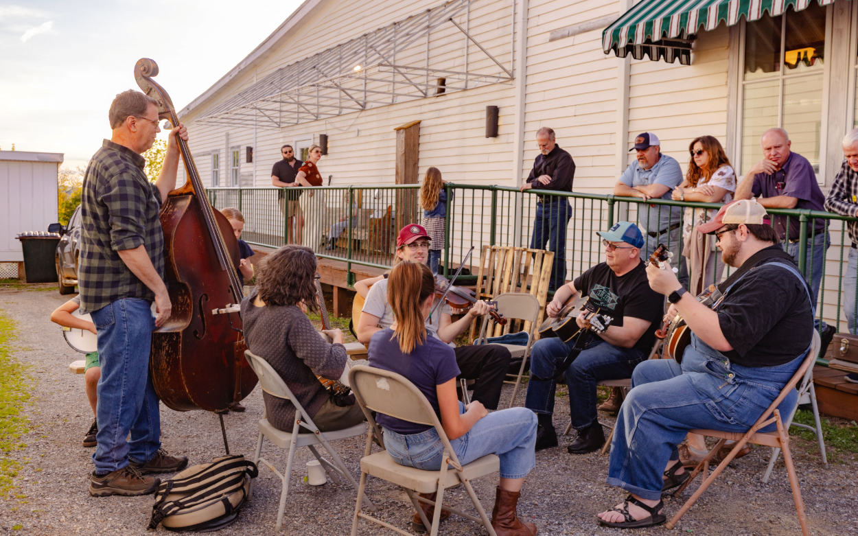 A group of people sit outside the Floyd Country Store to play music. They hold a variety of instruments, including guitars, fiddles, banjos and a stand up bass. 