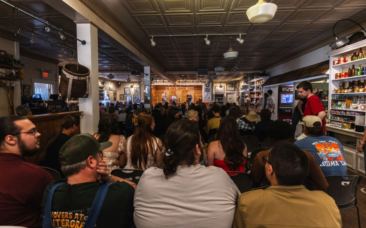 Students sit inside the dimly lit Floyd Country Store facing the stage, watching people perform bluegrass music. 