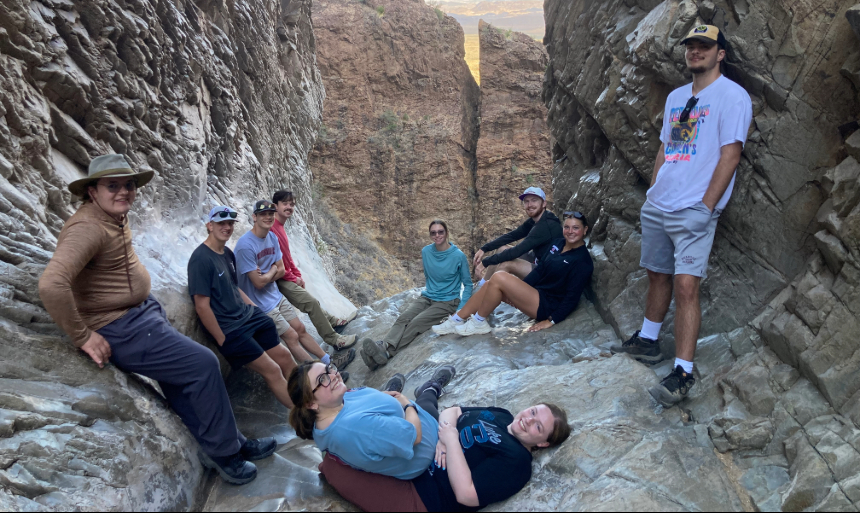 Ten people pose for a photo in between two cliff walls inside a national park. Some are sitting and others are standing, and in the foreground, one person lays on a rock while another sits on top of them. In the background, another gap between two cliff walls is visible. 