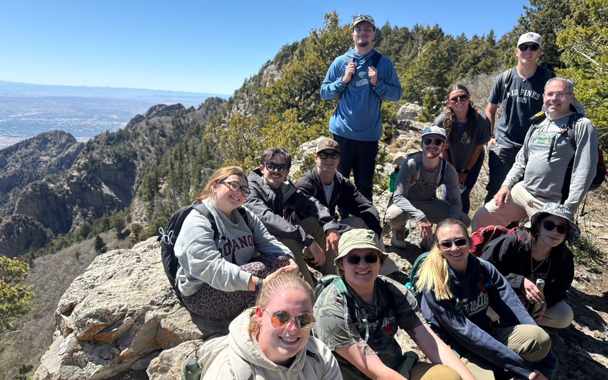 Twelve people pose for a photo on an overlook at a national park. Some are sitting on a rock while others are standing, and they are surrounded by greenery. 
