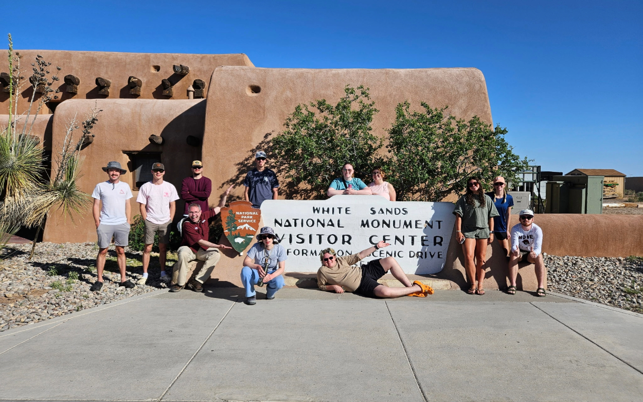 A group of students post with the White Sands National Monument Visitor Center sign. There is a short clay-colored building behind them, rocks and desert-type greenery.