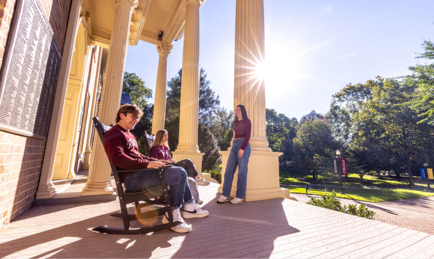 Students relax on the front porch of a historic building, some in rocking chairs and one leaning against a white column.