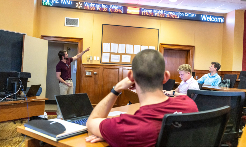 Students sit in a classroom listening to a speaker who is pointing at a stock ticker winding its way across the top of one wall of the classroom.
