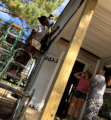 A student on a ladder doing work on the house while other students spot her