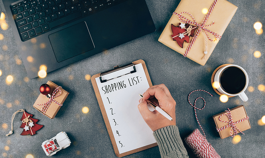 computer and gifts are seen with a hand writing a shopping list for gifts