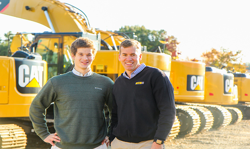 Ted Melnik and Dan Strelka stand in front of a line of heavy machinery, bulldozers, etc. 