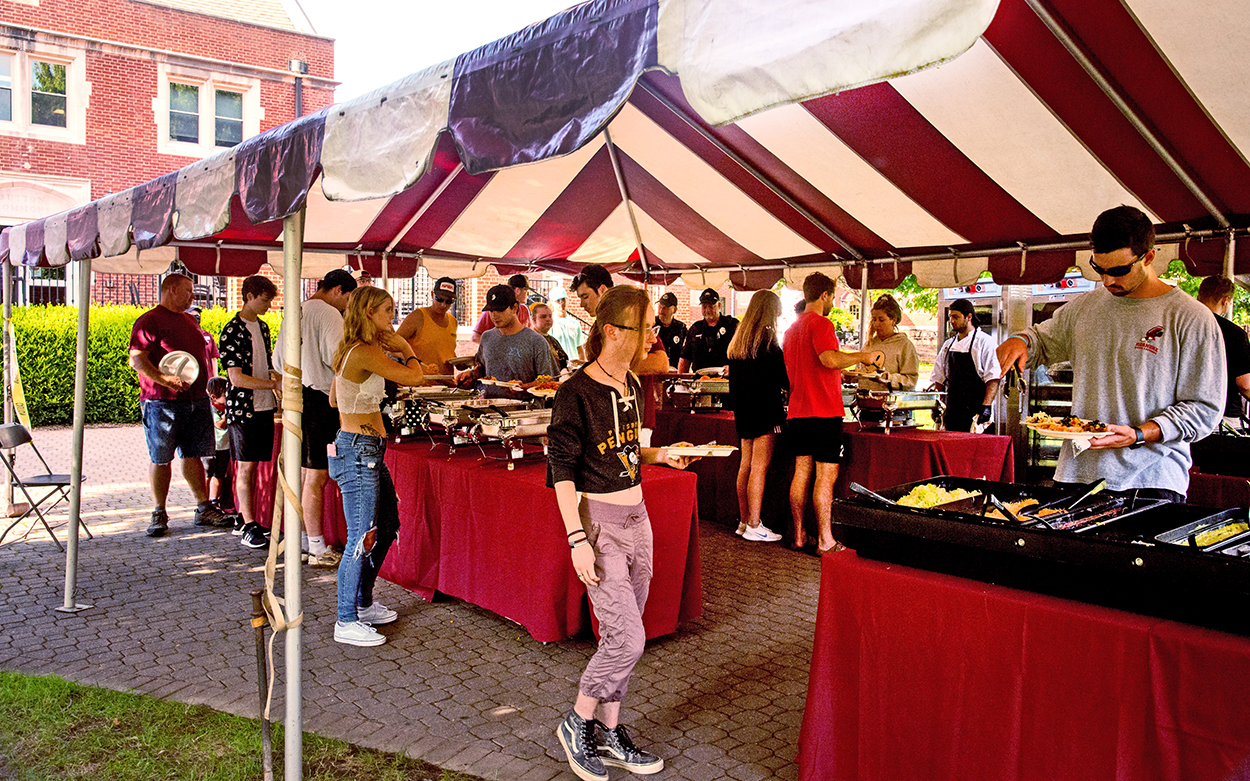 People serve themselves food from a buffet line under a red and white striped tent.