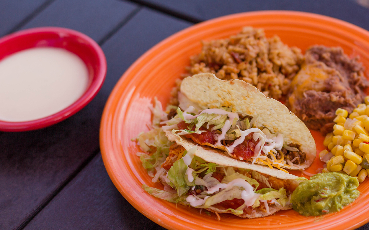 An orange plate with tacos and refried beans and guacamole and corn, with a side of queso sauce.