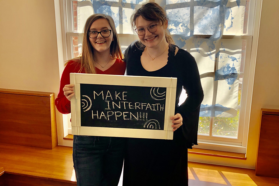 Two female students stand holding a chalkboard sign that says "Make Interfaith Happen!"