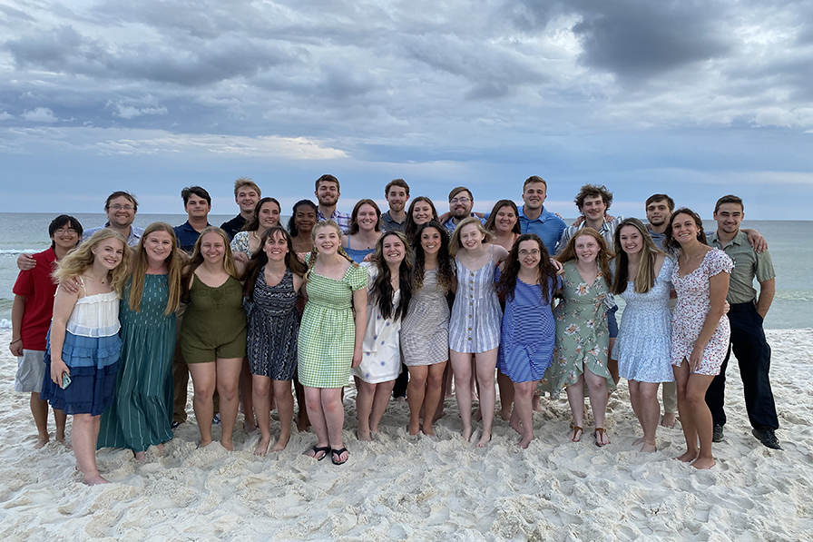 A huge group of young men and women pose together on a white sand beach with blue skies and puffy clouds in the background.