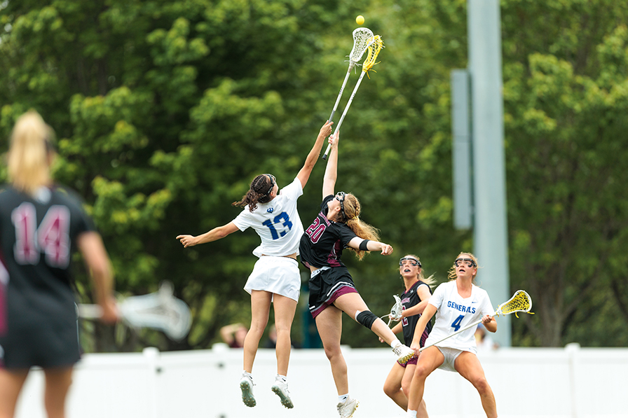 Two lacrosse players, one in a black Roanoke uniform and the other in a white uniform, jump up with their sticks in the air as they go for the ball at the same time.