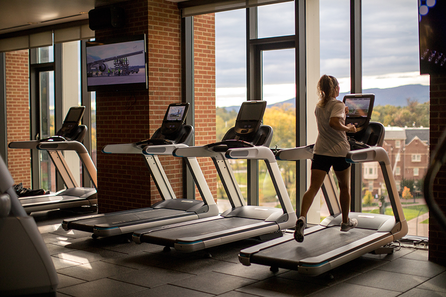 A young lady runs on a treadmill in front of a wall of windows, and outside you can see the brick dorms of campus.