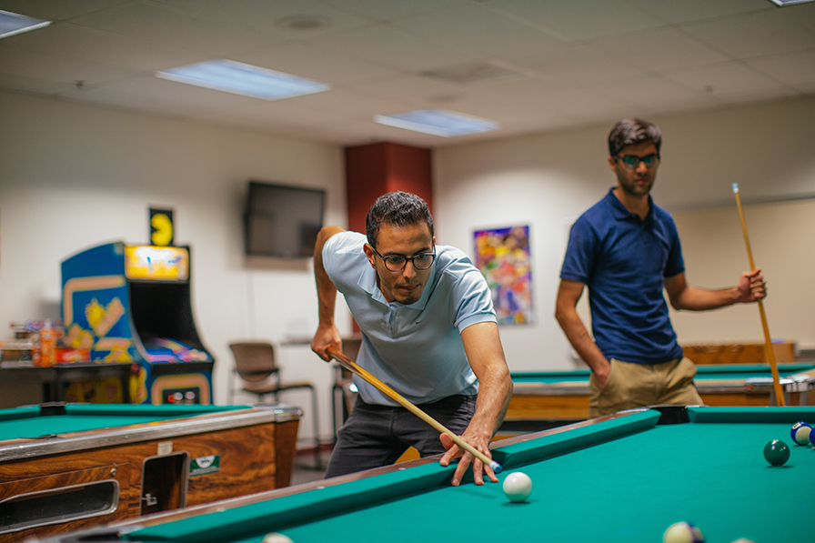 A young man leans over a green pool table to shoot while another guy looks on in the game room.