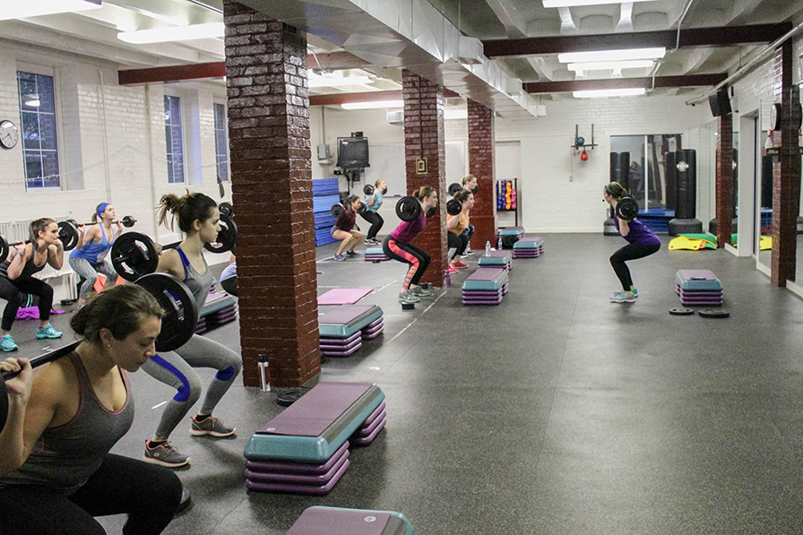 A group of women do squats with a barbell on their shoulders. One woman is in front of class facing them and instructing their movements.