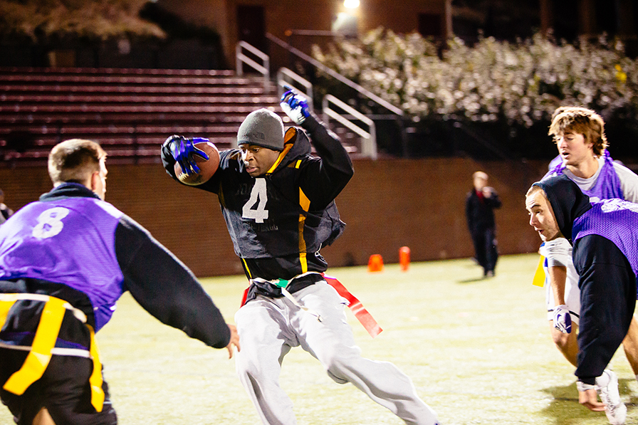 Two guys go for the flag on a third guy's belt in a game of flag football. They are outside on a field at night.