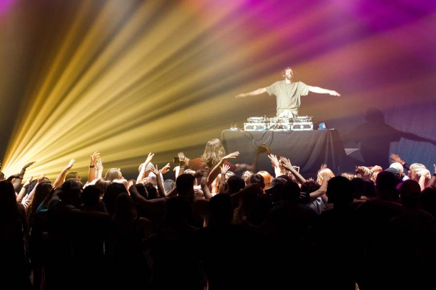 Performer stands on colorfully lit stage with audience in front of the stage