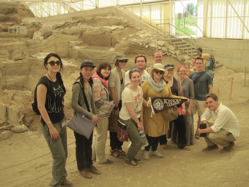Roanoke students and their professor hold up a Roanoke flag infront of a archeological dig