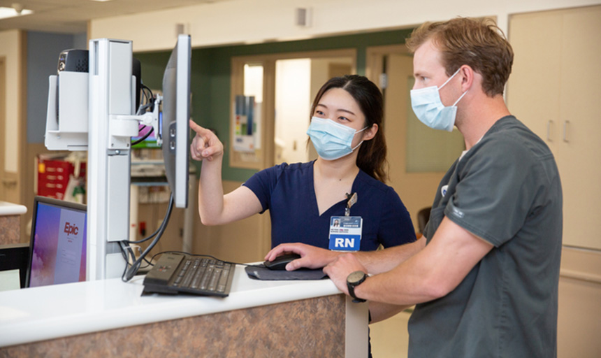 Two people look at a computer. they are wearing medical scrubs and masks and are in a medical setting.