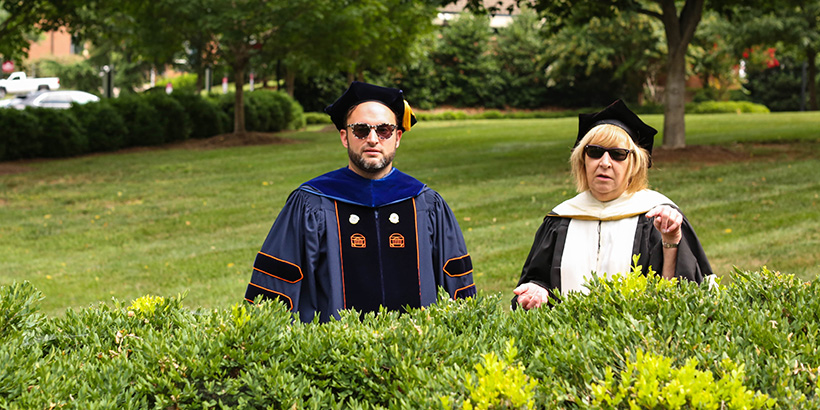 Two faculty members in academic robes, standing behind a shrub