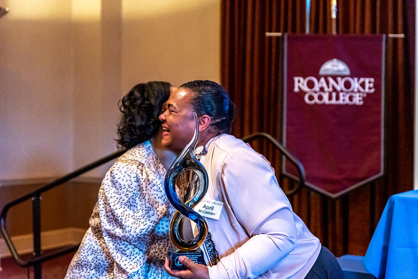 two women hug, one holding award in front of Roanoke College banner