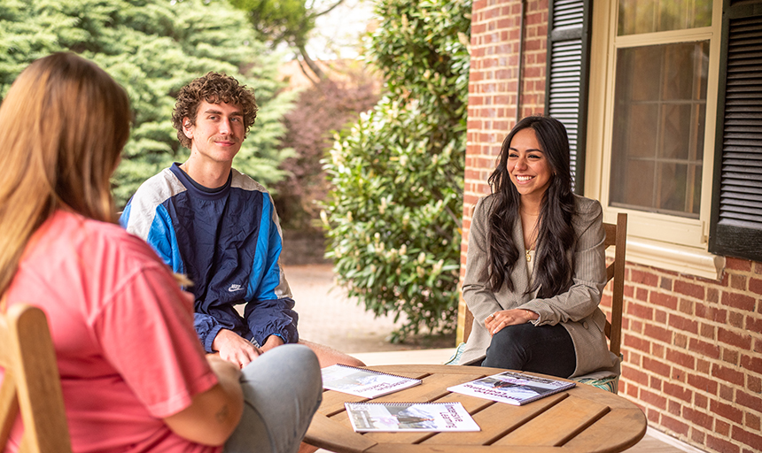 Counselor sitting at outdoor table with two students with green shrubs behind them.