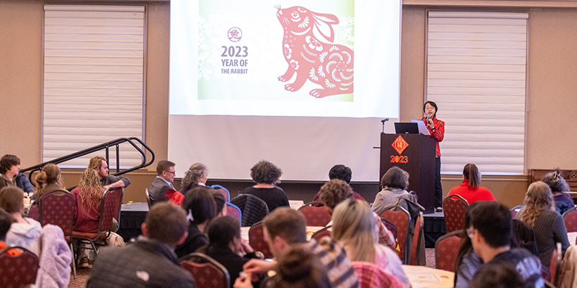 Professor Stella Xu stands at a podium to address a crowded audience gathered around tables in the Wortmann Ballroom