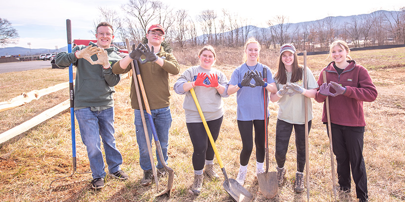 Students pose for picture while holding shovels and making hand symbols representing environmental fraternity Alpha Kappa Chi