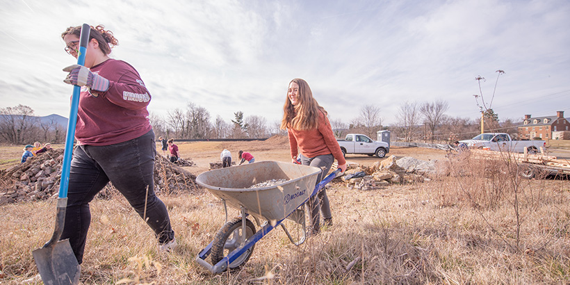 A student volunteer carries a shovel while another follows with a wheel barrow