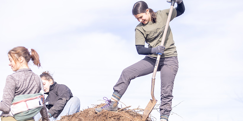 Student stands on pile of soil and digs out dirt with a shovel