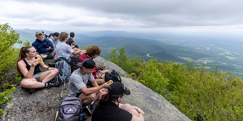 Students sit together on a mountain overlook for a lunch break of sandwiches and other food