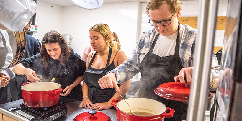 Three students dip into large, steaming pots of soup with spoons