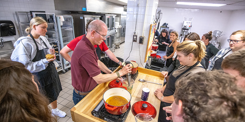 Students smile as they crowd around a table in a campus kitchen to watch Professor Adkins demonstrate a cooking technique