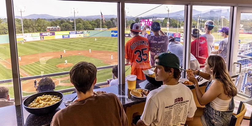 In the foreground, students inside a Salem Red Sox suite feast on hot dogs and other snacks while, in the background, more students gather around the railing of the suite's outside seating to watch the game on the baseball field below