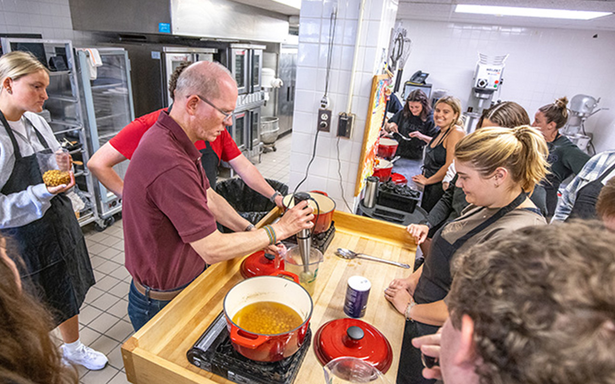 Students gather around a cooktop as their professor demonstrates a cooking technique